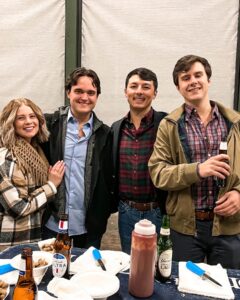 Four people stand smiling together indoors, dressed in casual winter clothing. Three men hold drinks, and a woman stands close to them. In front of them is a table with bottles, sauce, plates, and food.