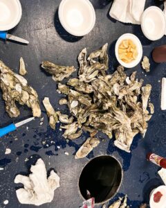 A tabletop covered in empty oyster shells, shucking tools, gloves, small bowls with lemon wedges, and napkins, suggesting an oyster shucking or seafood event.