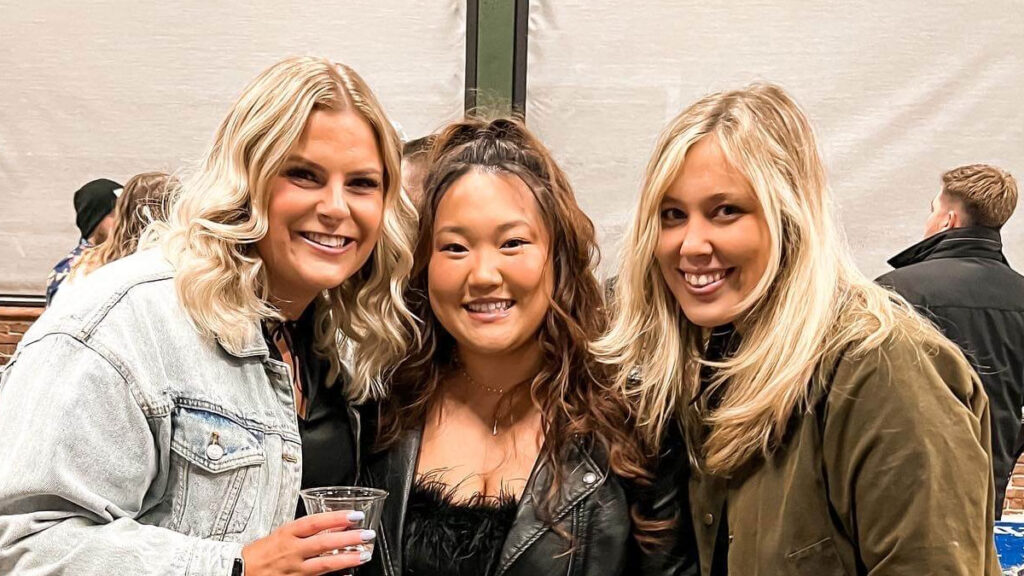Three women smile at the camera at an indoor event. The woman on the left holds a drink and wears a light denim jacket, the woman in the middle wears a black top, and the woman on the right wears an olive coat. People are in the background.