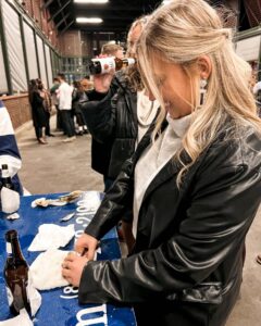A woman with blonde hair shucks oysters at a blue table in a crowded indoor event. She is smiling and wearing a black jacket. Other people and beer bottles are visible around her.