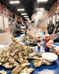 People stand around a long table covered with oyster shells, sauces, and plates, enjoying a communal oyster feast inside a large, industrial-style hall with steam rising from the oysters.