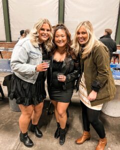 Three women pose together indoors at a casual social event. They are smiling and holding drinks, dressed in fashionable jackets and boots. The background shows other people and tables in a spacious, industrial-style setting.