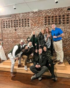 A group of eight adults pose and smile together on a wooden platform in front of a brick wall. Some are standing, some crouching, and a few are making peace signs. Casual attire and relaxed atmosphere.