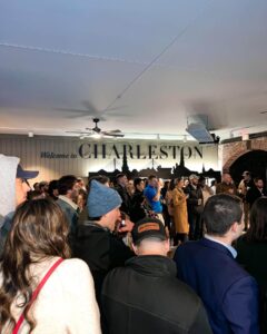 A crowd of people stands indoors, many facing a wall with a large sign that reads “Welcome to CHARLESTON.” The room has bright lighting and a visible ceiling fan.