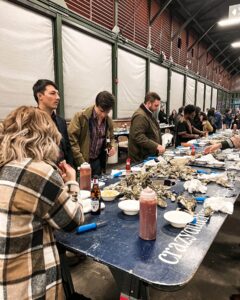 People stand and sit around a blue table covered with oysters, sauces, and condiments at an indoor oyster bar or seafood event. The atmosphere is lively, with several groups enjoying food together.