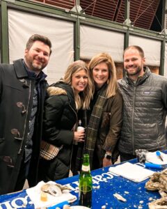 Four adults stand together smiling at an indoor event. They are dressed in winter coats and standing around a table with oysters, shells, and drinks. The background features a green and white structure.