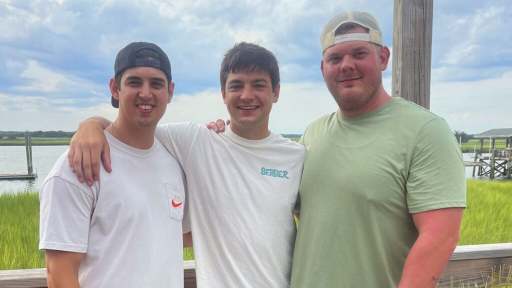 Three young men stand together outside by a wooden railing, smiling at the camera with a grassy marsh and water in the background under a cloudy sky.