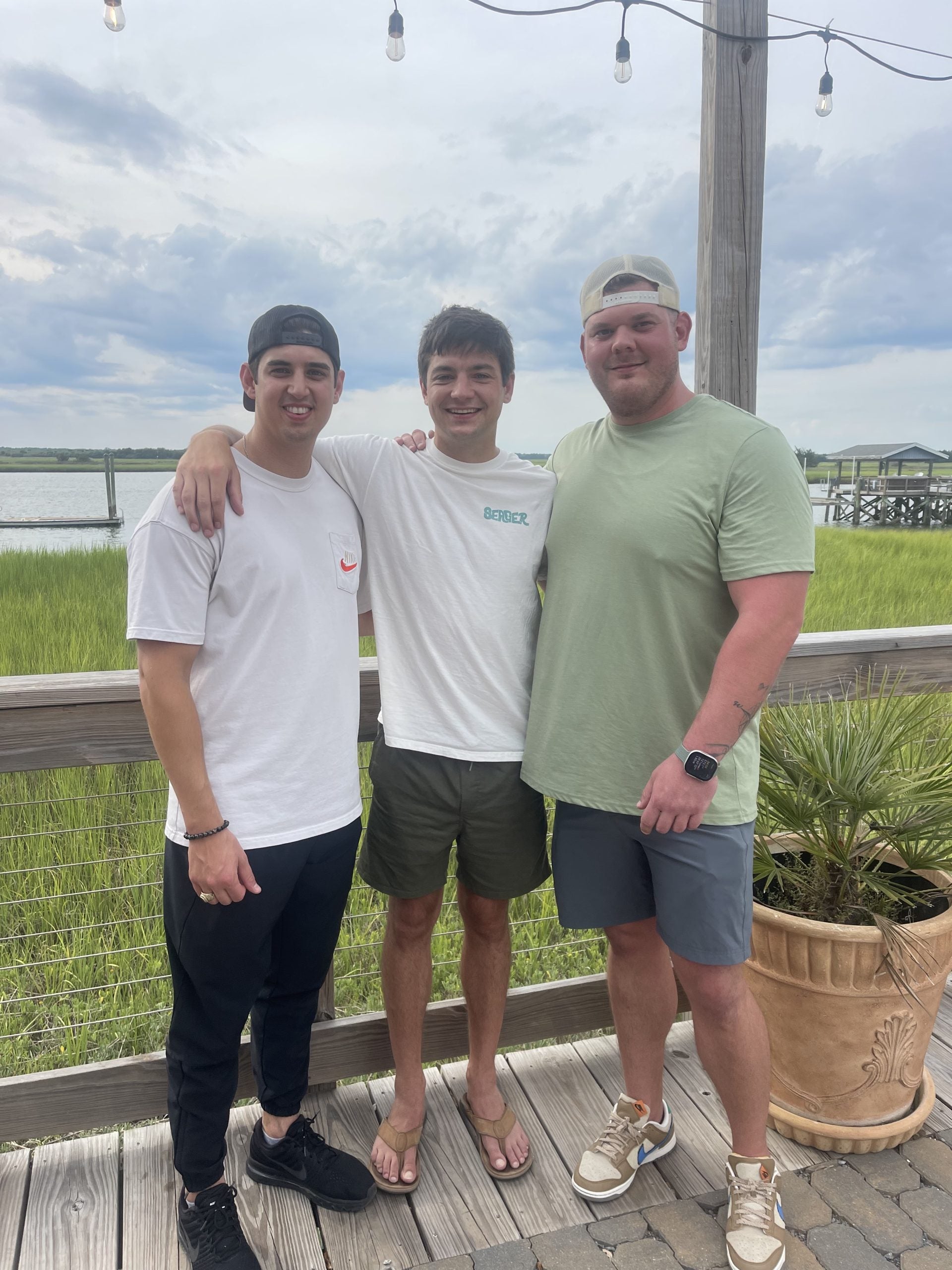 Three young men stand close together, smiling at the camera on a wooden deck with grassy marshland and water in the background under a cloudy sky. String lights are strung above and a potted plant is on the right.