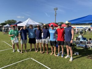 Eight men stand side by side on grass at an outdoor event, smiling at the camera. They wear casual summer clothes and hats. Several tents and people are visible in the background under a blue sky.