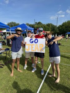 Five men stand outdoors on grass, smiling and posing with a handmade sign that says “GO JEAR” in blue and yellow letters. Some point at the sign; several wear hats and sunglasses. Blue tents and other people are visible in the background.