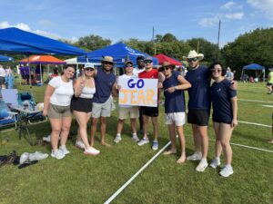Eight people stand on grass in front of colorful tents at an outdoor event, smiling and posing for a group photo. Two hold a handmade sign that says “GO JEAR” in large, bold letters.