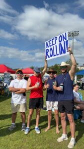 Four young men stand on grass at an outdoor event, smiling. One holds a blue and white sign reading “KEEP ON ROLLIN’!” They wear hats, sunglasses, and casual summer clothes. Other people, tents, and a light pole are in the background.