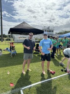 Two men stand barefoot on grass near bocce balls, smiling at the camera. One wears a dark shirt and shorts, the other a light blue Team Jean shirt and shorts. People gather under tents in the background on a sunny day.
