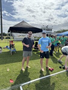 Two men stand barefoot on grass near bocce balls; one wears sunglasses and a dark shirt, the other wears a blue TEAM JEAN shirt and a cap. Tents, people, and a stadium are visible in the background.