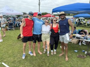Five people stand close together and smile for the camera at an outdoor event with tents and crowds in the background. They are dressed casually, and there is grass and scattered items on the ground around them.