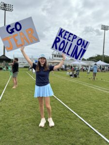 A young woman standing on a grassy sports field holds up two handmade signs reading GO JEAR and KEEP ON ROLLIN! She is smiling, wearing a blue cap, navy shirt, blue skirt, and sneakers. People and tents are visible in the background.