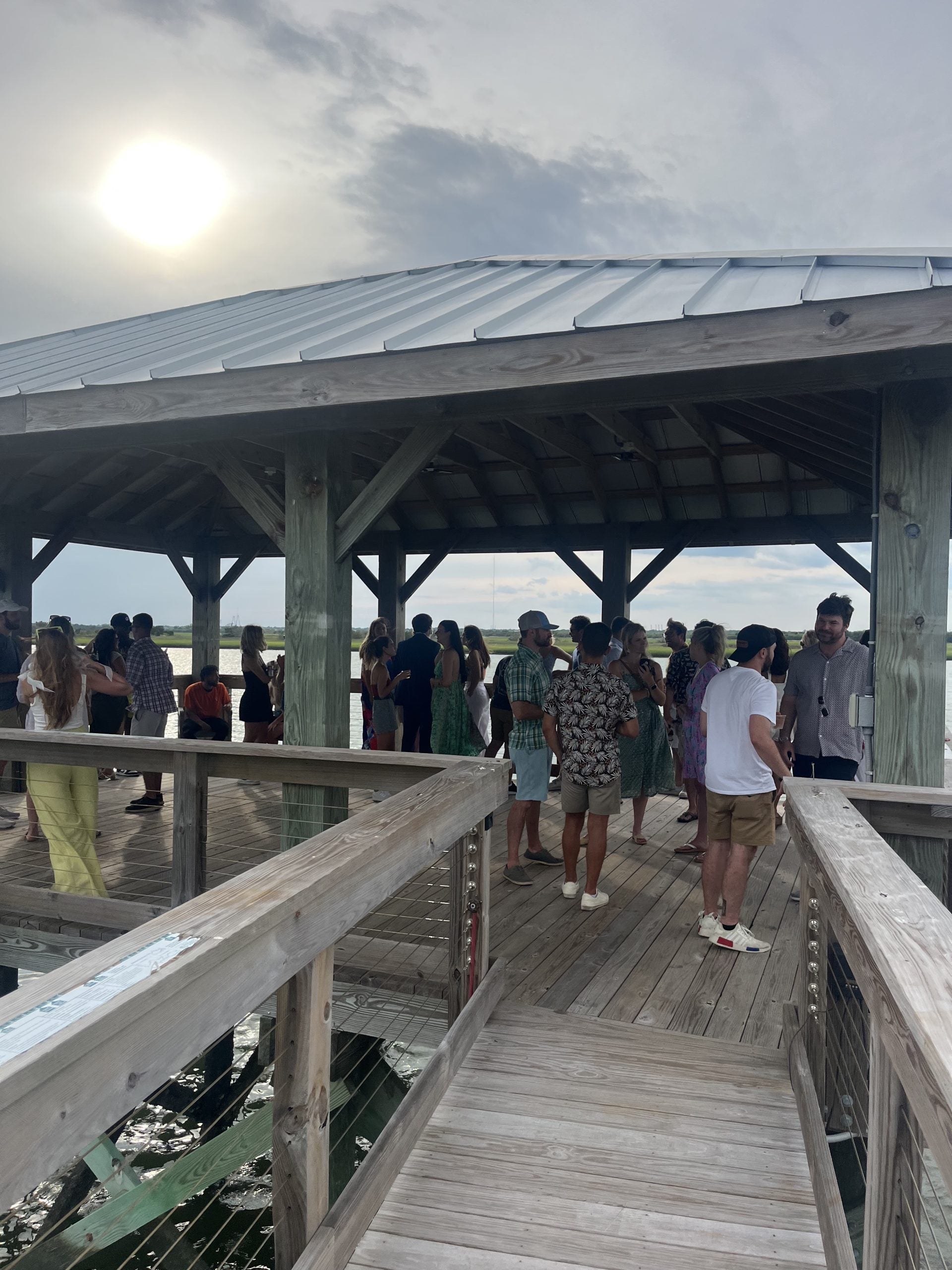 A group of people gather under a wooden pavilion on a dock over water, with the sun shining through a cloudy sky in the background. Some people are talking and others look out at the view.