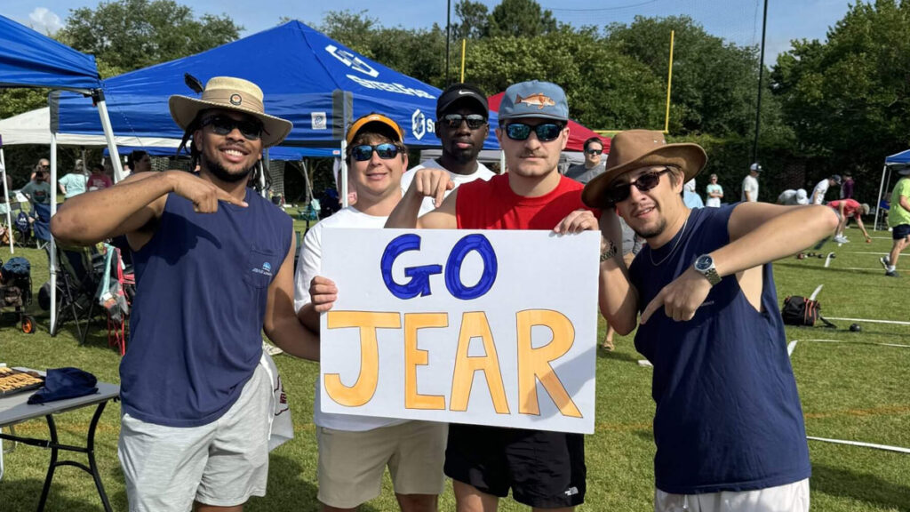 Five men stand outdoors on grass, smiling and pointing at a large white sign that says GO JEAR in blue and orange letters. Tents and people are visible in the background under a sunny sky.
