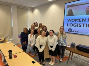 A group of twelve smiling women pose together in a classroom in front of a screen that reads “Women in JEAR Logistics.” Tables, chairs, and a laptop are visible in the room.
