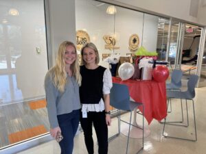 Two women smiling and posing together indoors near a tall table decorated with gift bags, balloons, and a red tablecloth. Office space with glass walls and award plaques is visible in the background.