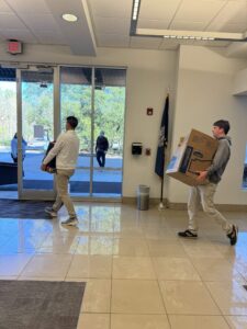 Two young men in khaki pants walk through a building lobby; one carries a large cardboard box. Sunlight streams through glass doors, and a person is visible outside. An American flag stands in the corner.