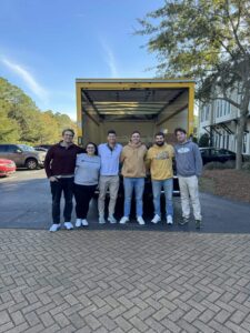 Six people stand side by side, smiling in front of an open yellow moving truck parked on a street with trees and buildings in the background.
