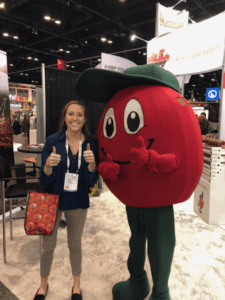 A smiling woman gives two thumbs up while standing next to a person in a large, red apple costume with a green cap and pants at a convention or trade show booth.