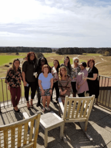 A group of ten women stand on a sunny balcony overlooking green fields and trees. Two wooden chairs and a small table are in the foreground. The women are smiling and dressed in casual, colorful clothing.