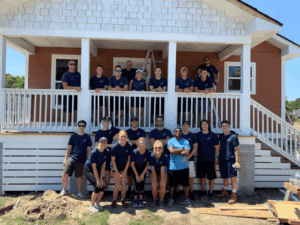 A group of people in matching navy shirts pose in front of a house under construction, with some standing on the porch and others on the ground, smiling for the photo on a sunny day.