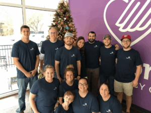 A group of twelve people, all wearing matching navy blue shirts, pose and smile together in front of a decorated Christmas tree and a purple wall with a large heart design.
