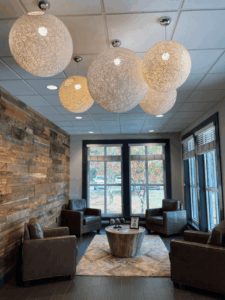 A cozy waiting area with four brown leather chairs around a round wooden coffee table, a rug, large windows, wood-paneled wall, and several spherical pendant lights hanging from a white ceiling.