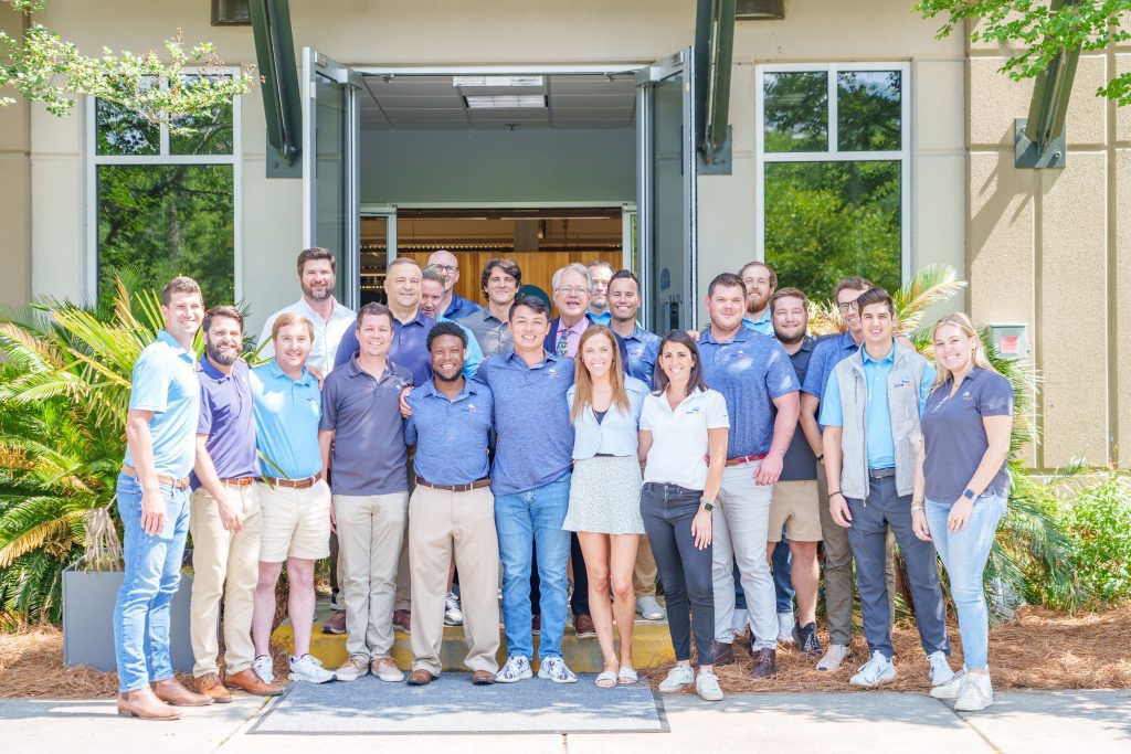 JEARLogistics-KeenEyeMarketing-038-1024x683 A group of 20 people, men and women, stand smiling together in front of a building entrance on a sunny day. Most wear casual summer clothing, and greenery surrounds the area—a perfect snapshot of team spirit at Why Work at JEAR.