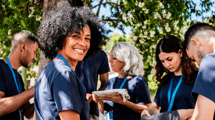 A group of people wearing navy blue JEAR Logistics shirts and name badges stand outdoors under a tree. One woman in front smiles at the camera, while others focus on their tasks as sunlight filters through the leaves above.