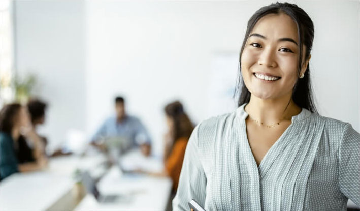 happy-customer A young woman with long dark hair smiling at the camera, standing in a bright office setting with a group of people working together in the blurred background.