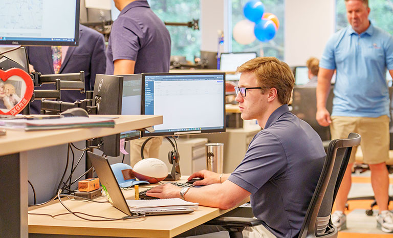 A man wearing glasses works at a dual-monitor computer in the busy, modern JEAR Logistics office. Other employees are present, including one walking in the background, with balloons and office supplies visible.