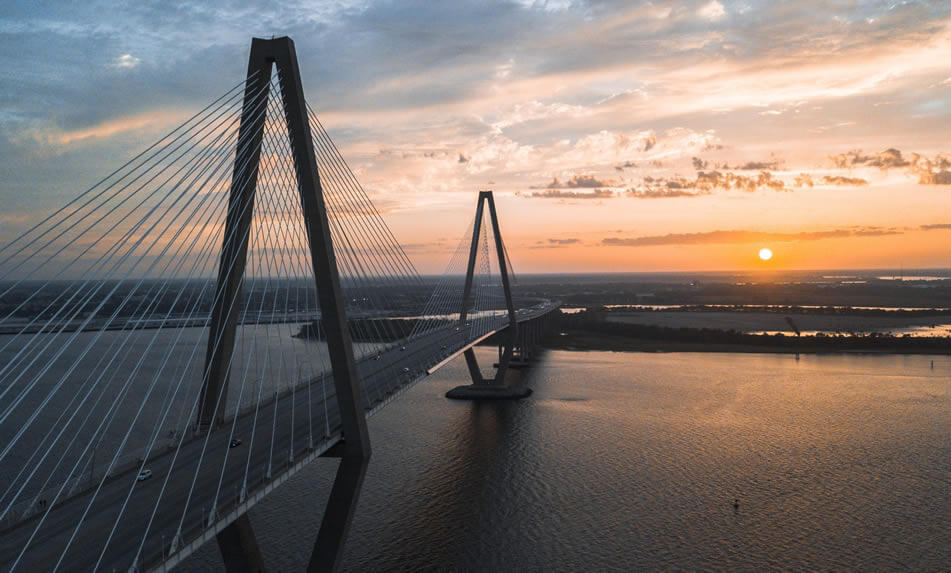 charleston-bridge A cable-stayed bridge stretches over a wide river at sunset, the sky adorned with clouds and the sun casting a warm glow—a scene reflecting JEAR Logistics Core Values of strength, reliability, and connection.