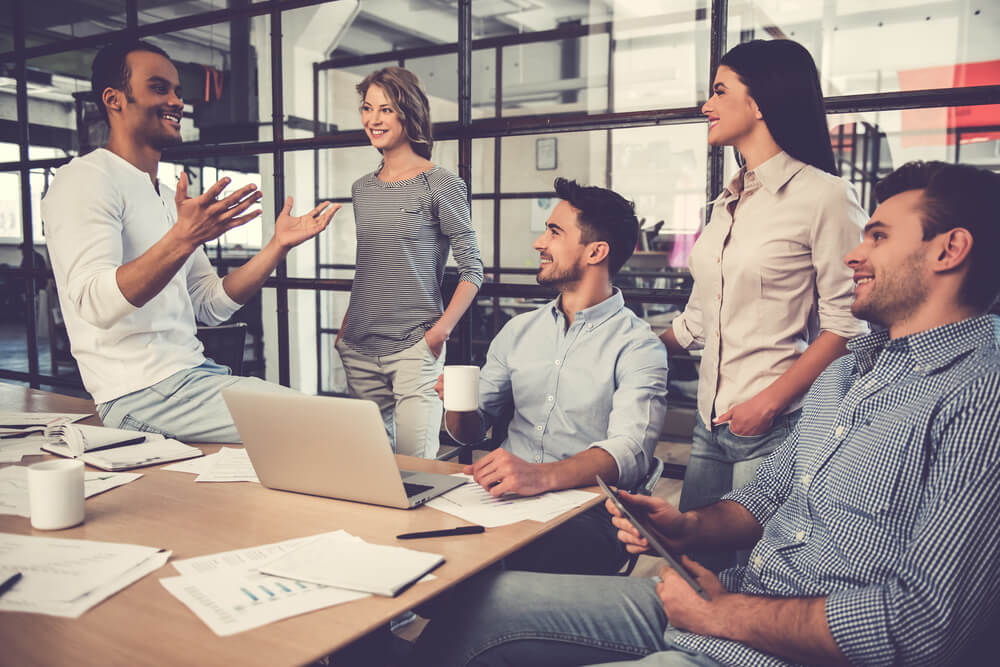 Five people are having a casual discussion in a modern office, reflecting JEAR Logistics Core Values. Two men sit at a table with laptops and papers, while three colleagues stand nearby, all smiling and engaged in conversation.