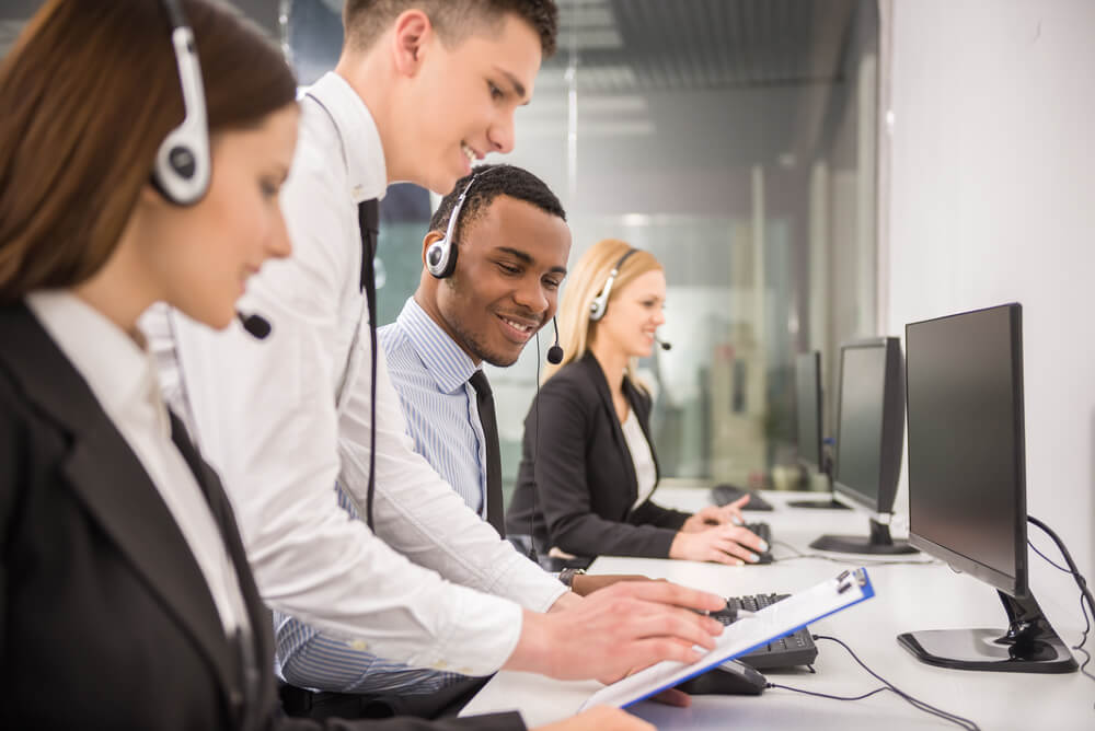 Four customer service representatives wearing headsets work at computers in an office, exemplifying JEAR Logistics Core Values. Two men in the foreground review notes on a clipboard, while a man and woman work at their desks in the background.