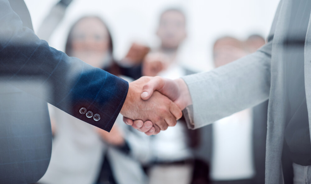 Two people in suits shaking hands, symbolizing a business agreement or partnership grounded in JEAR Logistics Core Values, with a group of blurred individuals standing in the background.