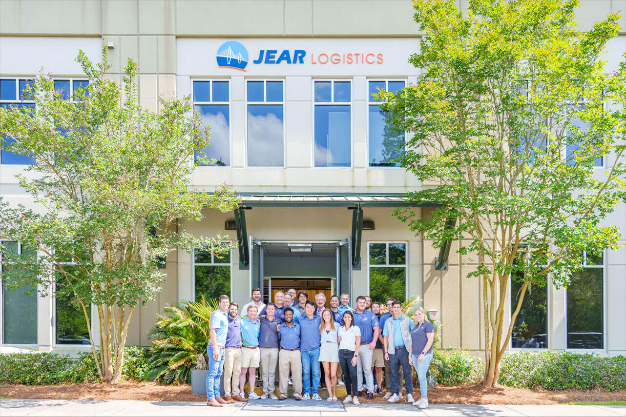 jear-building-team-photo A group of people stand smiling in front of a building with large windows and a sign that reads JEAR Logistics, embodying JEAR Logistics Core Values. Green trees and plants frame the entrance on a sunny day.