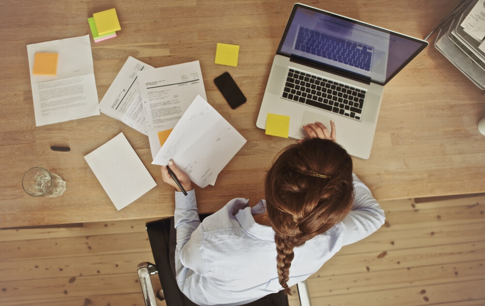 A woman with a braid works at a wooden desk with a laptop, documents, and notes, embodying JEAR Logistics Core Values as she writes on paper, focused and organized in her workspace, seen from above.