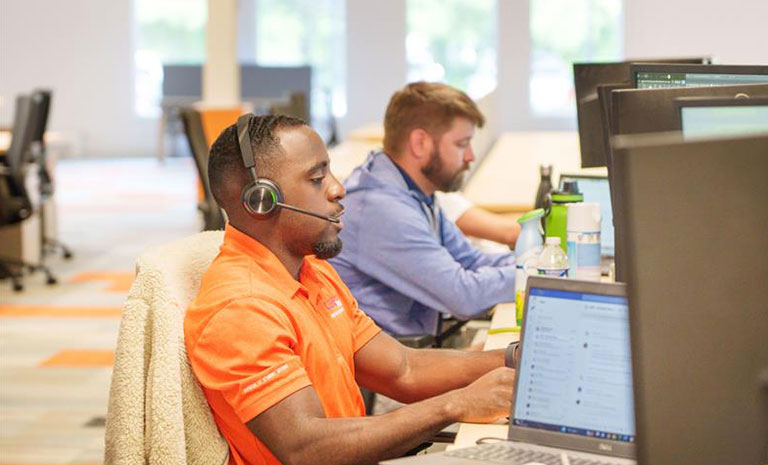 working-at-jear Two men sit at desks in an office, working on computers. The man in front wears an orange shirt and a headset, while the man behind him wears a blue shirt. Their focus highlights the professional environment that defines Why Work at JEAR.