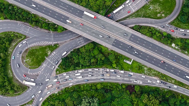 Aerial view of a busy highway interchange, vital for shipping and logistics, with overlapping roads, multiple lanes of traffic, and lush green trees and vegetation surrounding the area.
