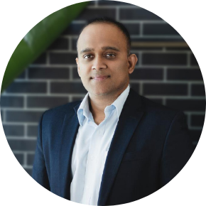 A man in a dark suit and white shirt stands in front of a wall with dark brick tiles, looking at the camera with a neutral expression.