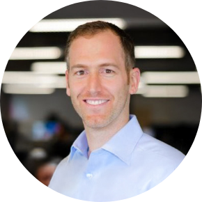 A man with short light brown hair, wearing a light blue collared shirt, smiles at the camera in an office setting with blurred lights in the background, reflecting a professional atmosphere in the shipping and logistics industry.