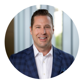 A man with short, brown hair wearing a blue plaid suit jacket and white dress shirt smiles at the camera; the background is blurred with vertical lines.