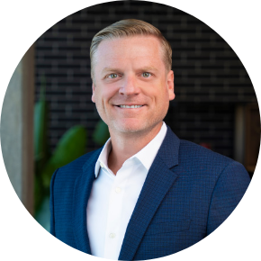 Smiling man with short blond hair wearing a blue suit jacket and white shirt, standing indoors with a dark brick wall and green plant in the background.