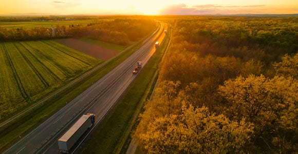 Aerial view of a highway surrounded by green fields and trees at sunset, with several trucks and cars traveling in both directions under a golden sky.