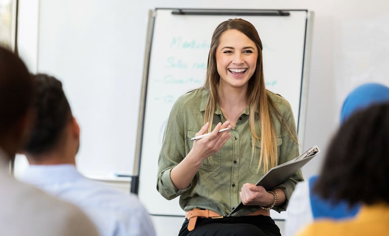 A woman stands in front of a group, smiling and holding a clipboard and pen, leading a discussion on Temperature-Controlled 3PL Shipping in a bright room with a whiteboard in the background.