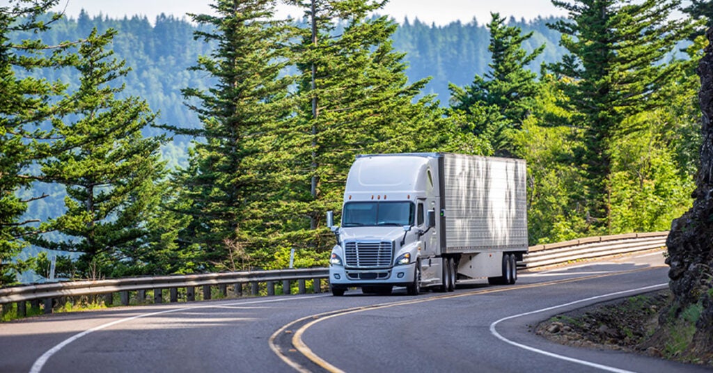 A white semi-truck drives on a winding road through a lush, forested area with tall green trees and distant hills, under a clear sky.